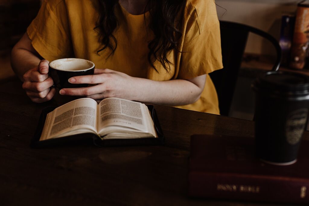 Girl, wearing a yellow tshirt, is holding a coffee mug with both hands with an open Bible on the table in front of her